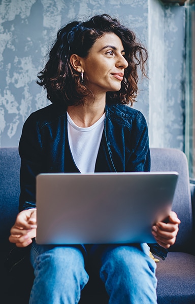 woman looks out window as she sits on couch with laptop on her knees