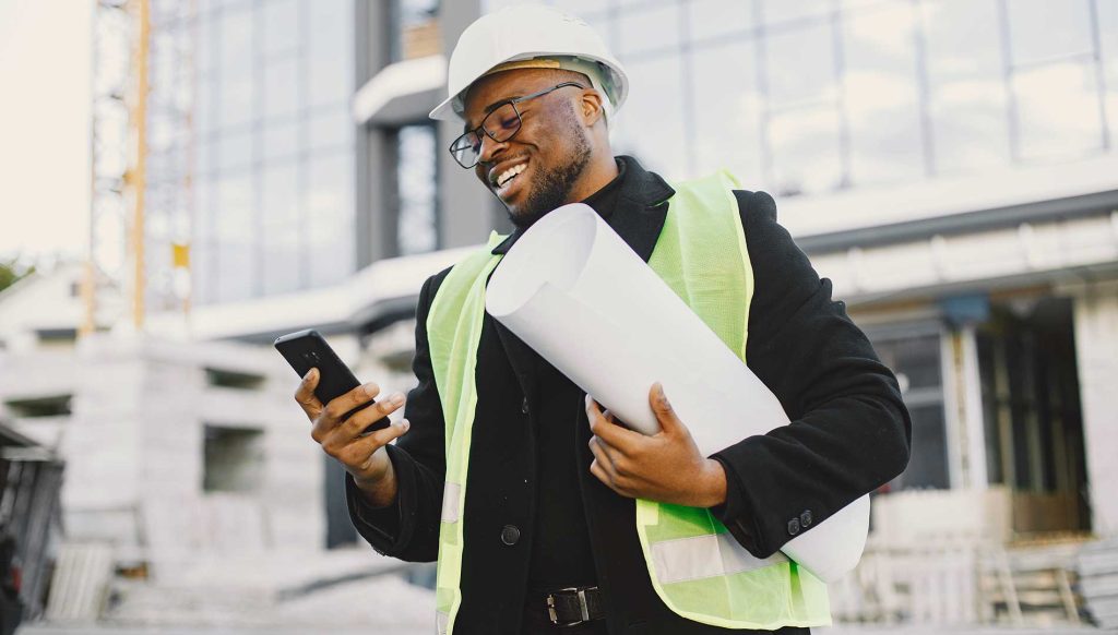 construction manager wearing hardhat holds rolled blueprints and texts on phone